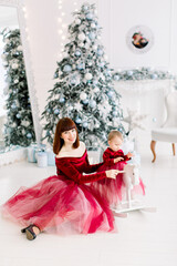 Happy family, woman mother and little girl playing near Christmas tree on Christmas eve at home. Little baby daughter swinging on white wooden horse. Christmas, New Year time