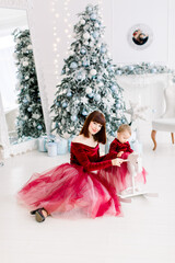 Happy family, woman mother and little girl playing near Christmas tree on Christmas eve at home. Little baby daughter swinging on white wooden horse. Christmas, New Year time