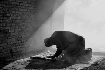 The silhouette of an Asian Muslim man, who is praying in a room with sunlight shining through the door of the mosque