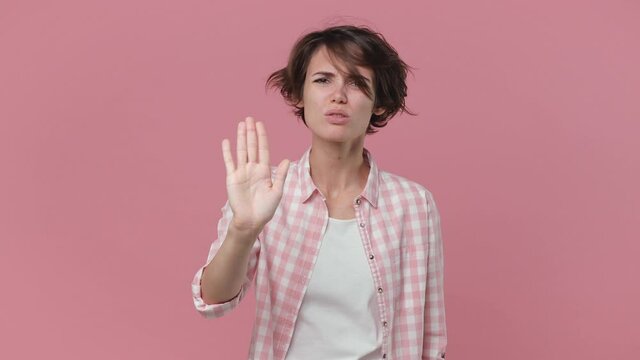 Displeased dissatisfied young woman 20s in basic checkered shirt posing isolated on pink background studio. People lifestyle concept. Looking camera showing stop gesture with palm crossed hands say no