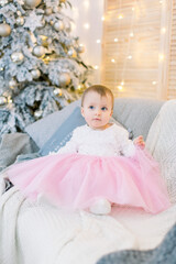 Little baby girl and Christmas decorations. Cute baby toddler girl, wearing in beautiful pink and white dress, sitting on stylish gray sofa, on the background of decorated Christmas tree