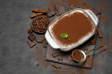 Traditional Italian Tiramisu dessert in glass baking dish and pieces of chocolate bar on concrete...