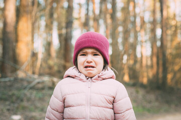 a 3-year-old girl in a pink jacket and a pink hat stands in the forest and cries 