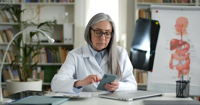 Elderly woman in glasses and white gown scrolling and touching smartphone sreen. Mature female doctor sitting at table in medical office. Concept of medicine and healthcare