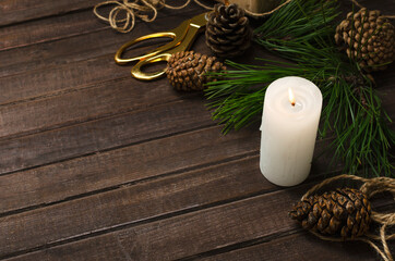 White candle and flame, cones, green fir branches, scissors and twine on the wooden table.Empty space