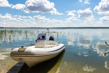 Big white modern fishing motorboat moored at lake or river shore sand pebble beach against scenic blue sky on bright sunny summer day. Inflatable boat parked on pond bank after water surface trip