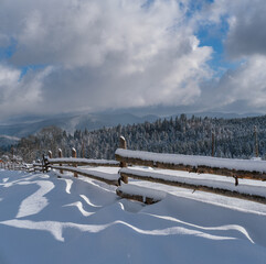 Picturesque waved shadows on snow from wood fence. Alpine mountain winter hamlet outskirts, snowy path, fir forest on far misty and cloudy hills.