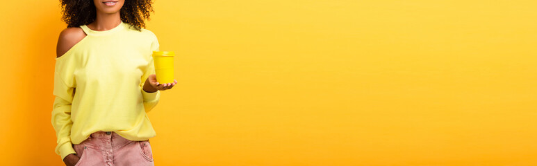 Obraz premium cropped view of african american woman holding reusable cup while standing with hand in pocket on yellow, banner