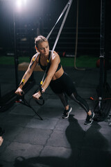 Fototapeta na wymiar Sporty young woman with perfect muscular body wearing black sportswear working out on simulator during sport training at modern fitness gym with dark interior.