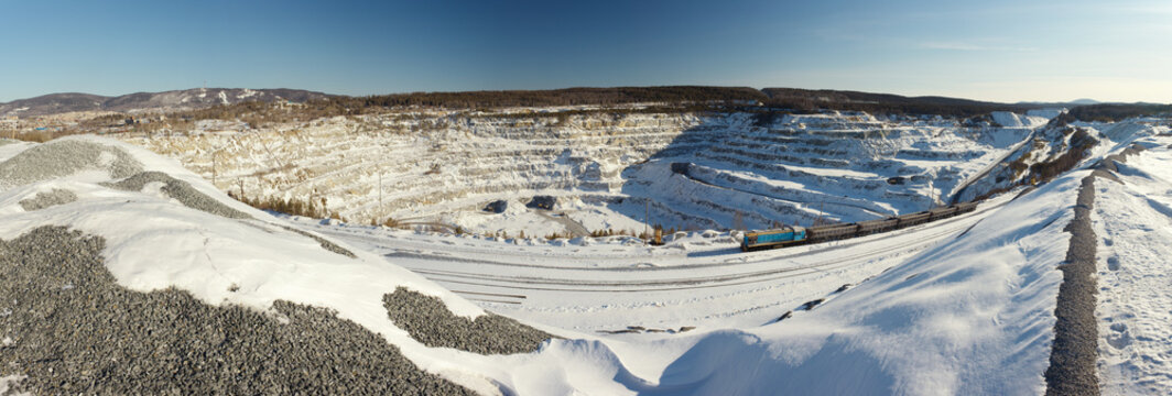 Wide-angle panorama of a huge stone quarry covered with snow in sunny weather.