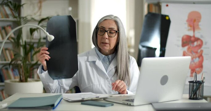 Female doctor showing x ray photo of chest while having online medical consultation. Mature lady in glasses and white rob using laptop while communicating with patient in her office