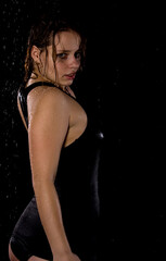 A beautiful girl in a black T-shirt stands under drops of water. Studio with water. Dark background. A beautiful model.