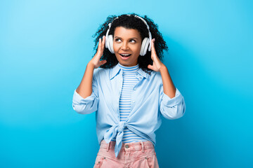 excited african american woman listening music in headphones on blue