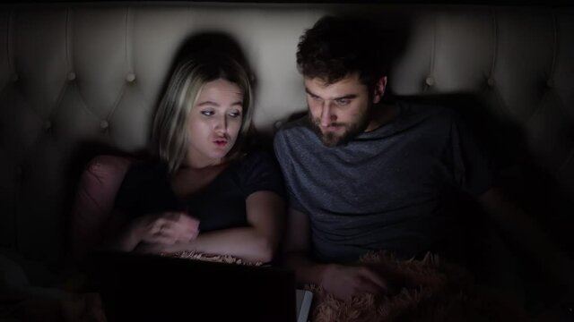 A man and a woman, a young couple are watching a movie on a laptop in bed before going to bed and they are attracted by a mobile phone call