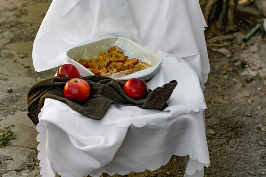 Collapsed soft baked apples in a white ceramic square shape on a special package. A white tablecloth is laid carelessly on the table, next to a dark napkin and ripe juicy red apples