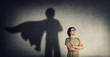 Confident young man keeps arms crossed smiling optimistic as casting a superhero with cape shadow on the wall. Motivated and ambitious guy tends to achieve success. Hero leadership and power concept - obrazy, fototapety, plakaty