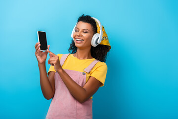 happy african american woman in headphones listening music and pointing with finger at smartphone with blank screen on blue