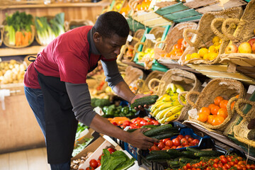 Confident African American seller in apron arranging fresh vegetables and fruits on greengrocery...