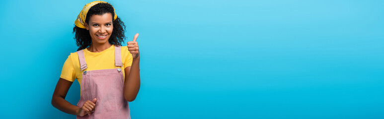 cheerful african american woman showing thumb up on blue, banner