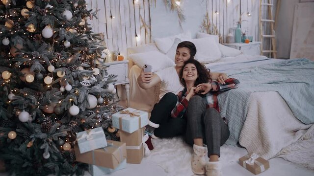 Young couple hugging and taking pictures with the phone, while sitting next to the Christmas tree at home. Bright living room decorated with bright Christmas tree lights. Happy winter holidays and