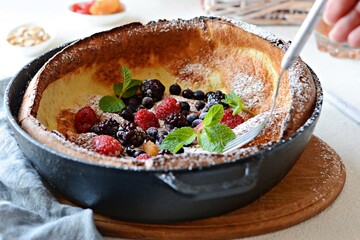 Fresh homemade dutch baby pancake with powdered sugar, fresh berries and mint for breakfast with a glass of milk and apple juice on a light background. Delicious sweet breakfast in a sunny interior. 