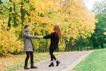 Young red haired woman puts on face mask while walking with young man in autumn park.