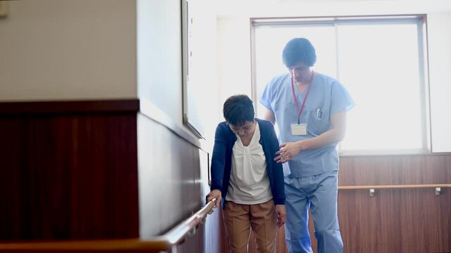 手すりにつかまりながら歩く高齢女性の介助をする介護士