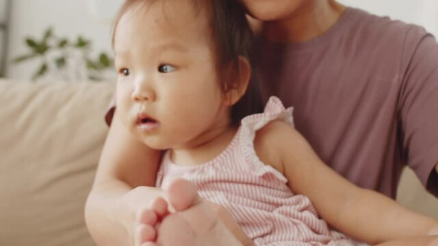 Tilt up close up shot of happy Asian mother sitting with sweet baby daughter on arms, kissing and touching her tiny feet while bonding at home