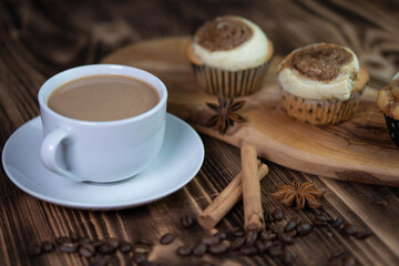 A white cup of coffee with homemade cinnamon cupcakes on a wooden board on a textured brown table composition closeup 