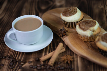 A white cup of coffee with homemade cinnamon cupcakes on a wooden board on a textured brown table composition closeup 