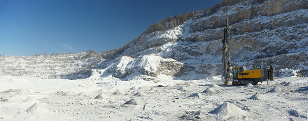 Mobile drilling machine in the snow-covered stone quarry in a winter sunny day, panorama.