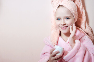 Obraz na płótnie Canvas a little girl in a pink dressing gown with a towel wrapped around her head smears her mother's cream on her face. pink background