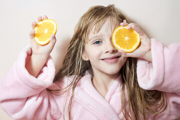 a charming blonde girl with wet hair in a pink bath robe covers her eyes with oranges on a pink background. one eye is open