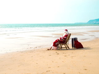 Santa Claus sits on a chair barefoot with a suitcase and a red bag with gifts resting on the seashore