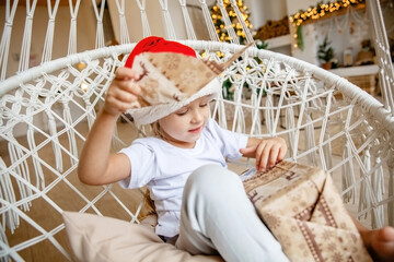 A four-year-old girl in a Santa hat sits in a wicker chair against the background of a Christmas tree and unwraps a gift