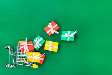 Small cart with scattered gifts on a green background. Red, green and yellow gifts with white bows in a shopping trolley. Christmas and Xmas gift concept. Copy space. View from above.