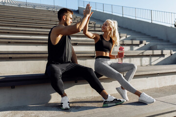 Fototapeta na wymiar Sportive young couple, man and woman sitting on stairs outdoors and giving each other high five, fitness people after successful workout