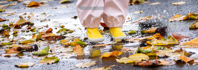A young child with yellow boots splashes in a rain puddle with autumn leaves