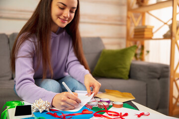 Fototapeta premium Comfortable. Woman writing letter, greeting card for New Year and Christmas 2021 for friends or family sitting at home with laptop. Holidays, celebration. Looks cheerful, happy, careful.
