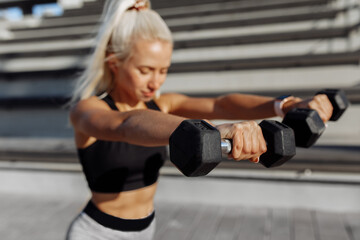 Attractive sportive young woman in sportswear stretching with dumbbells during warm-up outdoors on city stairs