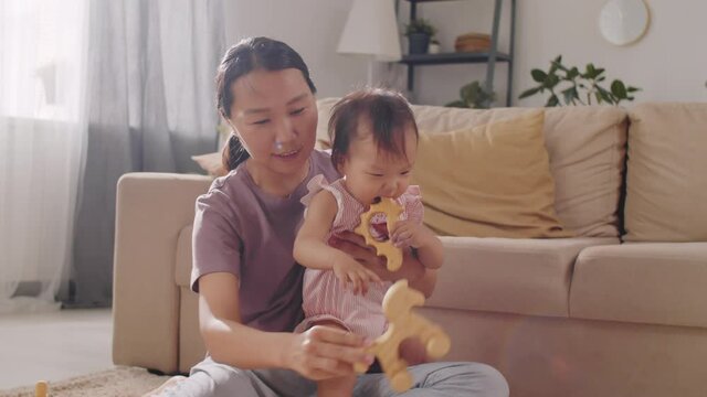 Cute Asian baby girl sitting with mother on the floor in living room and chewing plastic toy while playing together at home