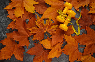 Two yellow dumbbells with tape measure (or measuring tape) on autumn leaves. Gym flat lay composition, healthy fitness lifestyle fall concept.