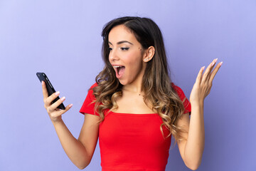 Young caucasian woman isolated on purple background looking at the camera while using the mobile with surprised expression