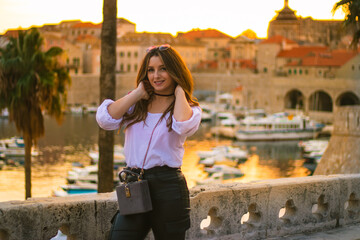 Fashionable attractive brunette posing on the city walls of Dubrovnik city as the sun sets during the golden hour. Small harbour in the distance illuminated by beautiful bright orange light