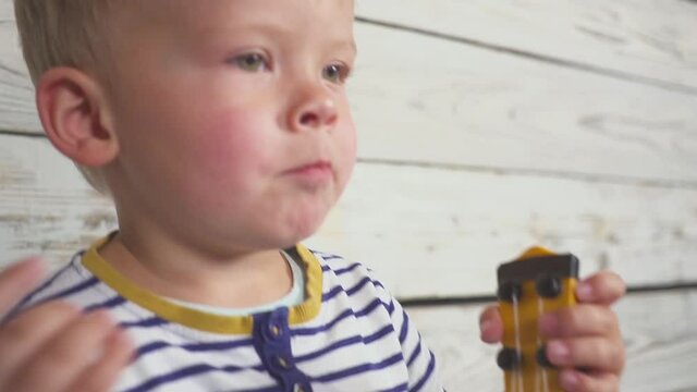 happy two year old boy plays his guitar or ukulele and sings songs, sitting in the wooden room.
