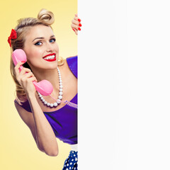 Woman with phone, in pin-up style dress, showing blank empty sign board with copy space area, over yellow background. Pinup blond girl in retro fashion and vintage studio concept.