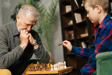 Boy playing chess with grandfather in chess game sitting at chessboard. Pensive beard grandfather...