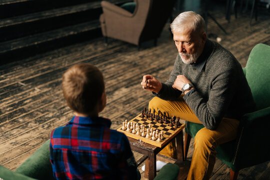Top view of aged-male grandfather playing chess with grandson sitting at desk in cozy dark living room with an authentic aristocratic interior. Grandpa tells little kid boy about the tactics of game.