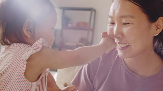 Joyous Asian woman smiling and speaking to adorable baby daughter while girl trying to feed mom during mealtime at home