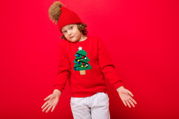 cool blond kid in warm hat and sweater with christmas tree on red background fooling around, christmas concept